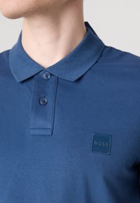 Kék polo póló texturált anyagból, állógallérral, két gombbal és négyzet alakú logopatch-csel, amelyen a "BOSS" felirat található a bal mellen.