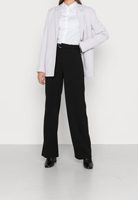 Heller Lavendelblazer, weißes Hemd mit Knöpfen, hoch geschnittene schwarze weite Hose, schwarze Stiefel mit Absatz. Glatter Stoff, taillierte Passform.