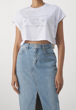 Femme portant un t-shirt blanc court avec des découpes en forme d'œil et d'étoile et une jupe en jean bleu clair taille haute avec une fente sur le devant.