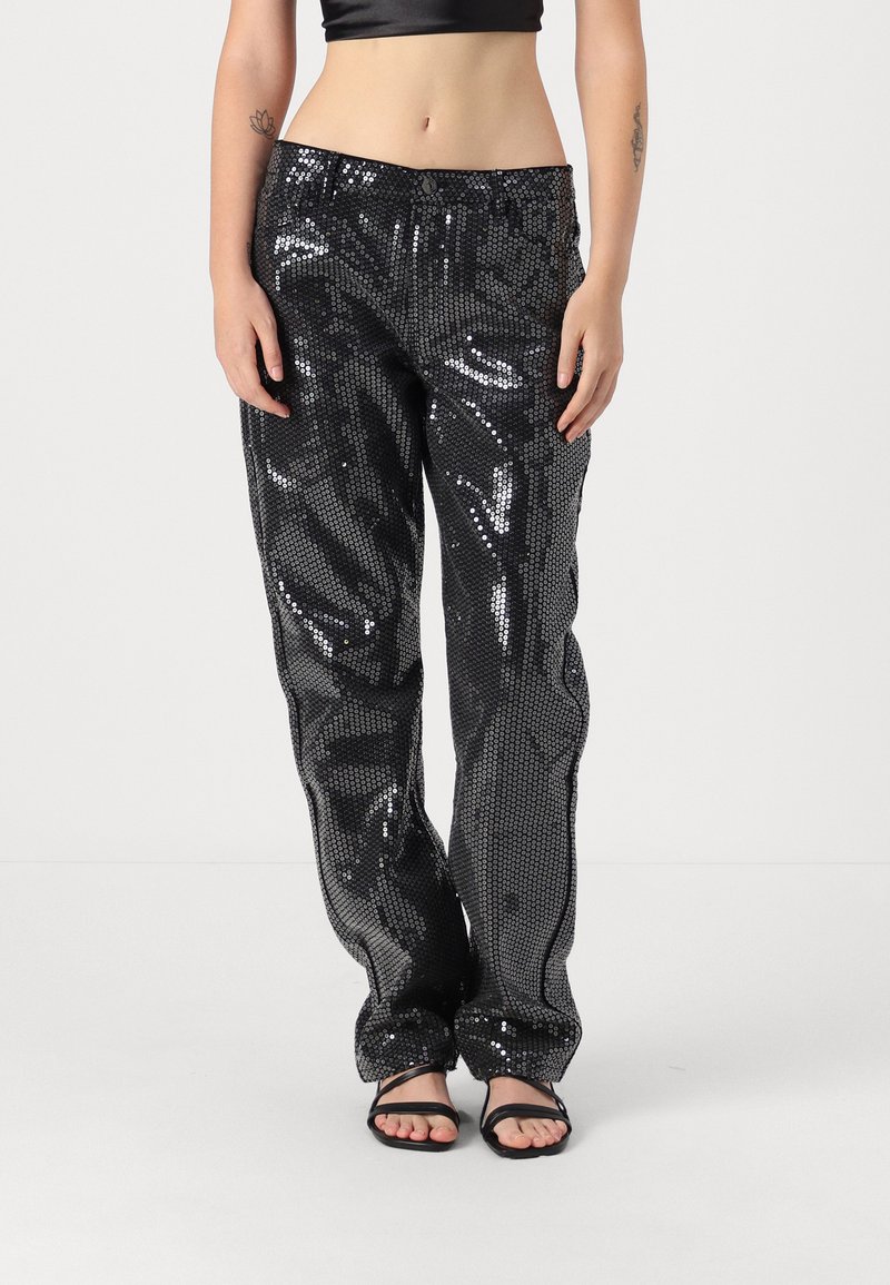 Pantalons noirs en sequins avec une coupe ample, présentant une texture brillante et un motif subtil. Associés à des sandales noires à brides.