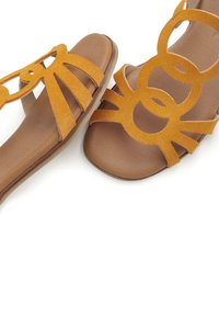 Gelbe Wildledersandalen mit flacher Sohle, die mit ausgeschnittenen kreisförmigen und linearen Mustern verziert sind. Das braune Innenfutter ergänzt die lebendige Farbe.