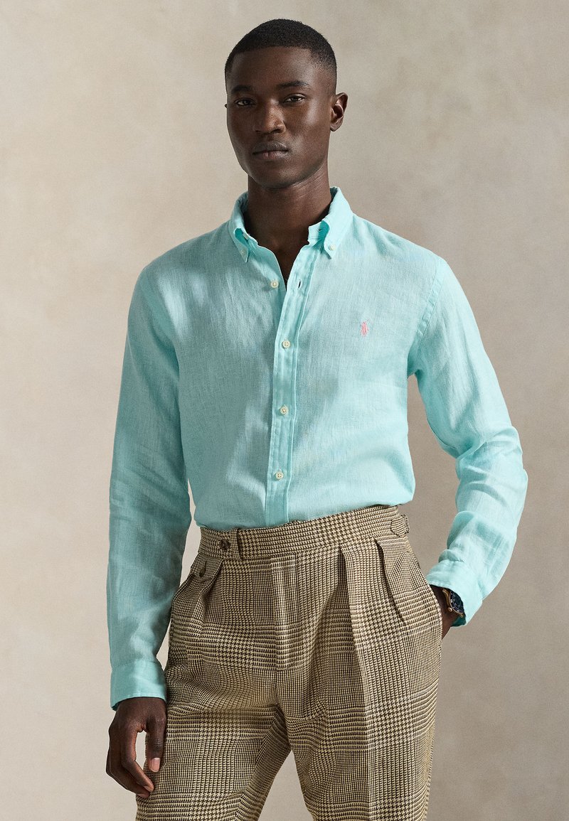 Camisa de lino azul claro con cuello abotonado y sutil branding, combinada con pantalones marrones de cuadros. Textura suave, corte casual.