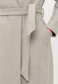Cappotto di lana grigio chiaro con una cintura in tessuto legata in vita e una mano infilata nella tasca destra.