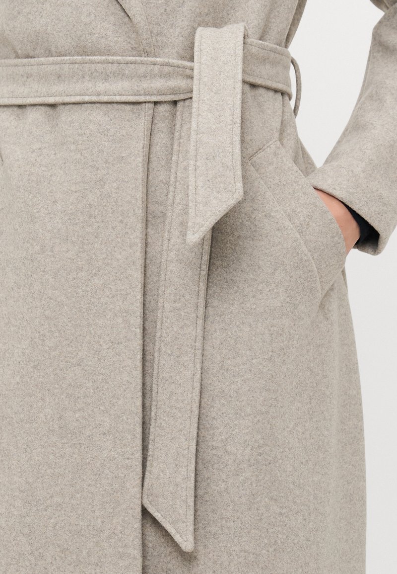 Manteau en laine gris clair avec une ceinture en tissu nouée à la taille et une main glissée dans la poche droite.