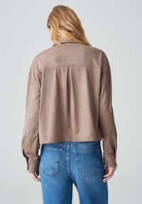 Cropped, brown suede jacket with a pleated back, long sleeves, and a classic collar, paired with blue denim jeans. Smooth texture.