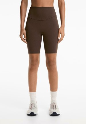 Shorts de vélo marron taille haute en tissu extensible, avec une texture lisse et une coupe ajustée, associés à des chaussures de sport légères et des chaussettes.