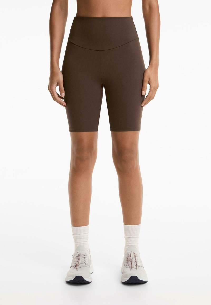 Brown high-waisted bike shorts made of stretchy fabric, featuring a smooth texture and a fitted design, paired with light athletic shoes and socks.