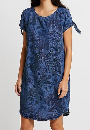 Kvinde iført en kortærmet blå kjole med mørkt blomsterprint og bundne ærmekanter, stående foran en ensfarvet baggrund.