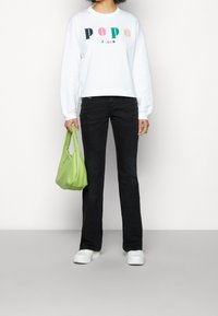 Persona vistiendo un sudadera blanca con el colorido texto "Pepe Jeans", jeans negros, zapatillas blancas y sosteniendo un bolso de mano verde lima.