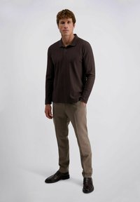 Dunkelbraunes Langarm-Polohemd mit Kragen und zwei Knöpfen. Kombiniert mit hellbraunen gemusterten Hosen und dunklen Slippern. Neutrale Hintergrund.
