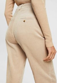 Beige corduroy pants with back buttoned pocket worn with a matching ribbed long-sleeve top, hand relaxed by side.