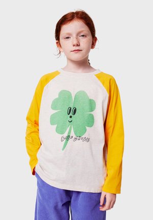 Langarm-T-Shirt mit einem cremefarbenen Körper, gelben Raglanärmeln, einem grünen Kleeblatt-Graphic und schwarzem Text. Weicher Stoff und lockere Passform.