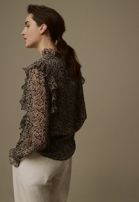 Sort floral chiffon bluse med ruffled detaljer langs ærmerne og kraven, parret med lyse bukser.