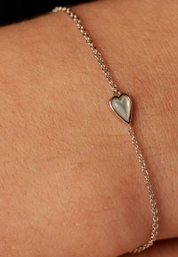 Silver chain bracelet featuring a small heart charm with a white, reflective surface, resting against skin. Simple, delicate design.