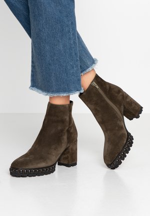 Kobieta nosząca ciemnooliwkowe zamszowe botki na kostkę z okrągłymi ćwiekami na podeszwie i słupkowymi obcasami, w połączeniu z niebieskimi dżinsami z postrzępionymi brzegami.