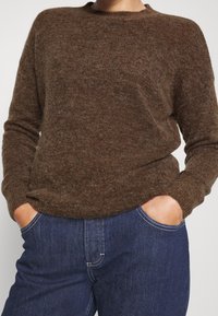 Personne portant un pull marron texturé légèrement rentré dans un jean bleu foncé, les mains dans les poches avant, sur un fond uni.