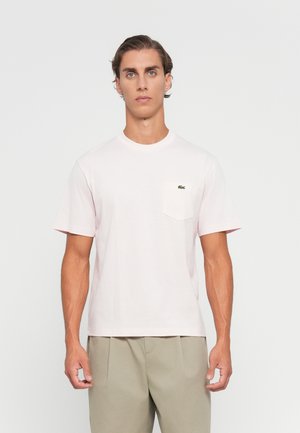 Joven con cabello corto castaño que lleva una camiseta de manga corta rosa claro con un bolsillo y pantalones beiges, de pie frente a un fondo blanco liso.