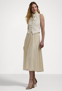 Colete de linho bege com botões frontais, combinado com uma saia longa pregueada bege com padrão de renda. Sandálias de salto em cetim completam o visual.