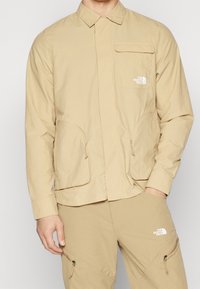 Beżowa lekka kurtka z szpiczastym kołnierzem, suwakiem z przodu, kieszenią na piersi oraz dwiema kieszeniami po bokach; z logo na lewej piersi.