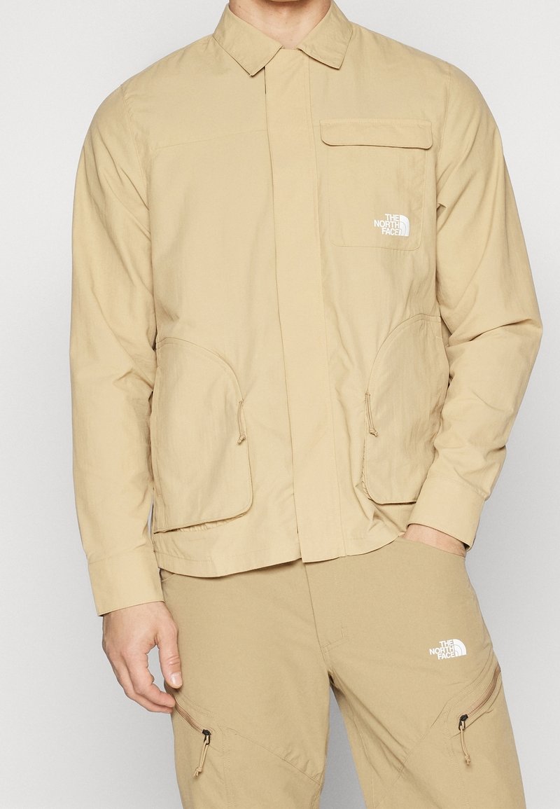 Beżowa lekka kurtka z szpiczastym kołnierzem, suwakiem z przodu, kieszenią na piersi oraz dwiema kieszeniami po bokach; z logo na lewej piersi.