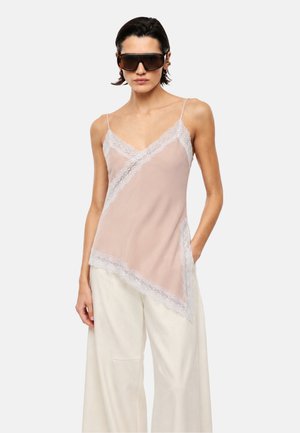 Donna con grandi occhiali da sole scuri, una canottiera rosa pallido con bordo in pizzo e pantaloni bianchi ampi, in piedi con una mano in tasca.