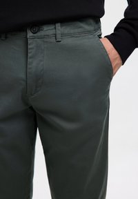 Person trägt dunkelgrüne Hose mit Hand in der Tasche und schwarzem Oberteil, sichtbar an der Taille vor hellem Hintergrund.