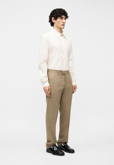 Άνδρας που φοράει κρεμ πουκάμισο με κουμπιά, μπεζ παντελόνι προσαρμοσμένης ραφής και μαύρα δερμάτινα παπούτσια, στέκεται μπροστά σε ένα απλό λευκό φόντο.