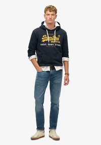 Marineblauer Hoodie mit Frontlogo, Kordelzugkapuze und Kängurutasche, kombiniert mit hellblauen Jeans und beigen Sneakers mit braunen Akzenten.