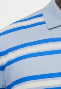 Ανοιχτό μπλε πλεκτό πόλο πουκάμισο με οριζόντιες μπλε και λευκές ρίγες στον ώμο και το γιακά εμφανείς.