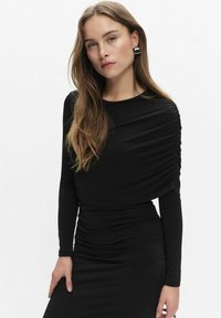 Czarna sukienka z długim rękawem, z marszczeniami na ramionach i bokach, wykonana z gładkiego materiału; w połączeniu z geometrycznymi srebrnymi kolczykami.