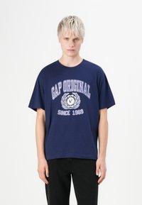 Sötétkék pamut póló rövid ujjal, levendula színű grafikai nyomattal, amely tartalmazza a "GAP ORIGINAL" és a "SINCE 1969" feliratokat.