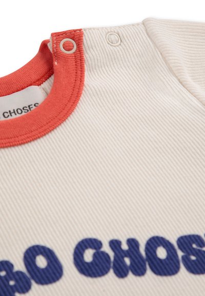Maglietta a coste color crema con colletto rosso, caratterizzata da una grande stampa in blu. La parte frontale presenta chiusure a scatto per una facile vestibilità.