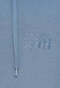 Blåt stof med en fletning af snørre og prægede "JACK & JONES"-logo på brystområdet af en hættetrøje eller sweatshirt.