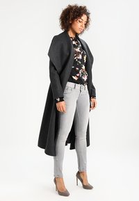 Un long manteau gris foncé, un chemisier floral, un jean slim gris clair et des talons gris. Le manteau a un design drapé ; le chemisier présente des motifs floraux colorés.