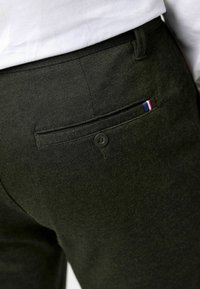 Gros plan sur un pantalon noir avec une poche arrière boutonnée et une petite étiquette en tissu rouge, blanc et bleu près de l'ouverture de la poche.