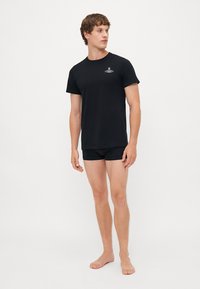 Black cotton t-shirt with short sleeves and a small logo on the chest, paired with black boxer shorts, worn by a standing male model.