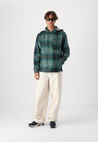 Grön och svart rutigt flanellskjorta med huva, knäppning och bröstficka; bärs med krämfärgade byxor och svarta sneakers.