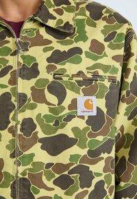 Rumena kamuflažna jakna z različnimi zelenimi in rjavimi vzorci, zadrgo in belim logotipom Carhartt na prsnem žepu.