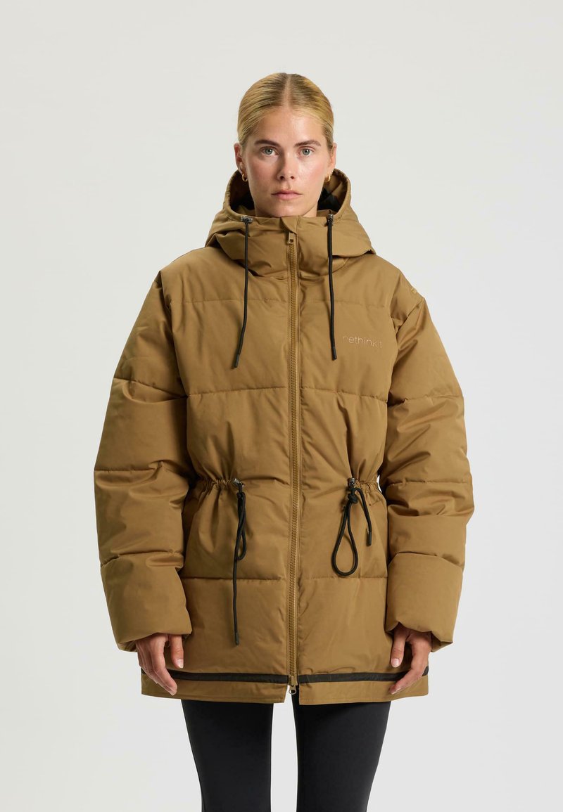 Vrouw in een lichtbruine gewatteerde jas met capuchon, zwarte koorden en rits, staand tegen een effen lichte achtergrond.