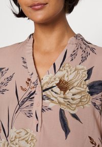 Blommig blus i mjuk rosa tyg med V-ringning, med stora krämfärgade och gyllene blommor, mörkgröna blad och rosa knappar.