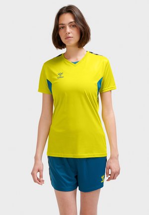 Ung kvinde iført gul kortærmet sportstrøje og blå shorts står mod en ensfarvet lys baggrund.