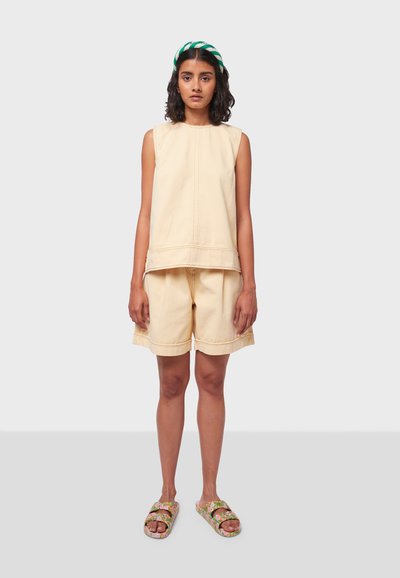 Top beige senza maniche con scollo rotondo e pantaloni corti abbinati, con cuciture visibili. Indossato con sandali a scivolo con motivo floreale.