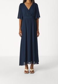 Mörkblå maxiklänning med fladdrande ärmar, V-ringning, åtsittande midja och flödande tyg. Bärs med ljusa, remmade sandaler.