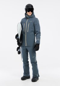 Männliches blaues Snowboard-Outfit mit einer wasserdichten Jacke mit Reißverschlusstaschen, passenden Hosen, Handschuhen und schwarzem Helm. Snowboard in der Hand gehalten.