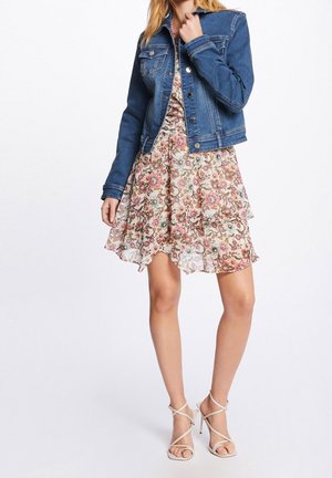 Kobieta w niebieskiej dżinsowej kurtce na floralnej sukience, w beżowych sandałach na wysokim obcasie, stojąca na białym tle.