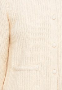 Flauschige Strickjacke in Off-White mit strukturiertem Stoff, runden Knöpfen und einer kleinen aufgesetzten Tasche mit geflochtenem Detail auf der Vorderseite.