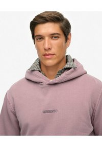 Sudadera de lavanda con capucha, con un logotipo de "SUPERDRY" bordado en lavanda más oscura. Debajo, se puede ver una camisa de franela a cuadros.