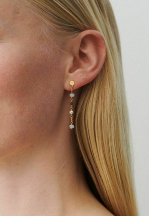 Krupan plan uha plave žene koja nosi zlatnu naušnicu s lancem, bijelim perlama i malom bisernom kuglicom.