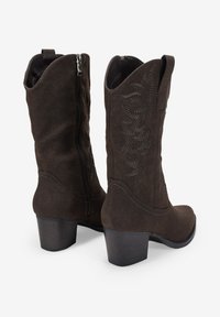 Botas de vaquero de ante sintético marrón con cremalleras laterales, punteras afiladas, tacones cuadrados y costuras detalladas a lo largo de los lados exteriores.