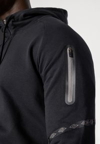 Čierna mikina s kapucňou s vreckom na zips na ľavom rukáve, hladkou textúrou a dekoratívnym vzorom na manžete.